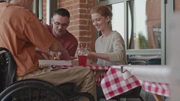 Μέτριο πλάνο του αναπηρικού αμαξιδίου νεαρός άνδρας και οι φίλοι του τρώνε πίτσα και κουβεντιάζουν σε υπαίθριο καλοκαιρινό καφέ την ημέρα - Πλάνα, βίντεο