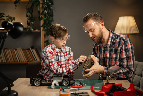 Vader en zoon in beschermende glazen met soldeerbout voor het repareren van speelgoed auto thuis in de avond. Het begrip familie en saamhorigheid. Kopieerruimte - Foto, afbeelding