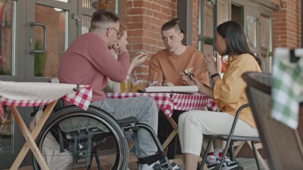 Молодий кавказький чоловік у інвалідному візку та його три друзі їдять піцу та спілкуються за столом у літньому кафе - Кадри, відео