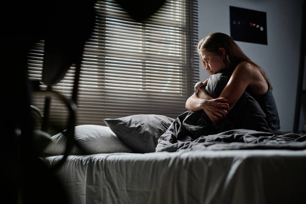 A zaklatott tinédzser lány oldalnézete, aki átöleli magát, miközben az ágyon ül, és a névtelen személy internetes zaklatására gondol. - Fotó, kép