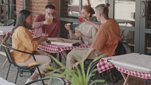 Gruppo di giovani diverse persone sedute a tavola in un caffè estivo all'aperto alzando bicchieri di plastica con limonata. Uno di uomo seduto in sedia a rotelle - Filmati, video