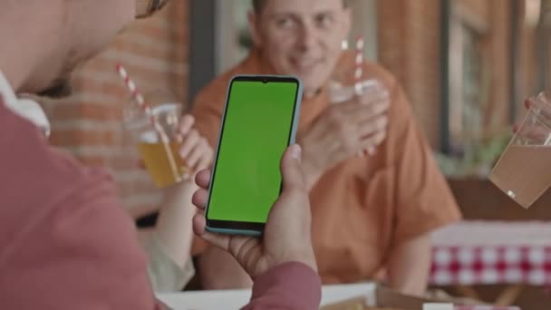 Olkapään yli kaveri tarkastellaan älypuhelin vihreä malli näytön syödessään yhdessä ystävien kanssa kesällä kahvilassa ulkona - Materiaali, video