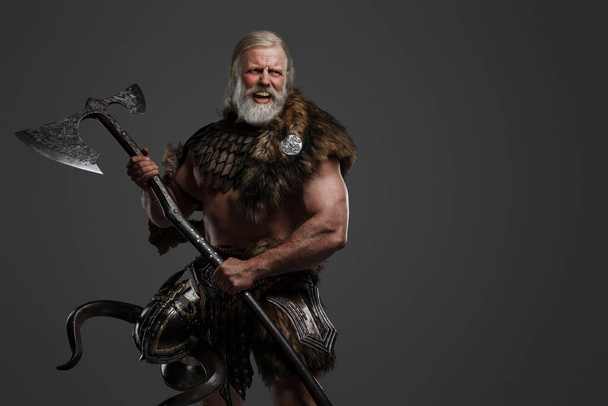 Un viking viking furieux, aux longs cheveux blancs et à la barbe, vêtu de fourrure et d'armure légère sur fond gris - Photo, image