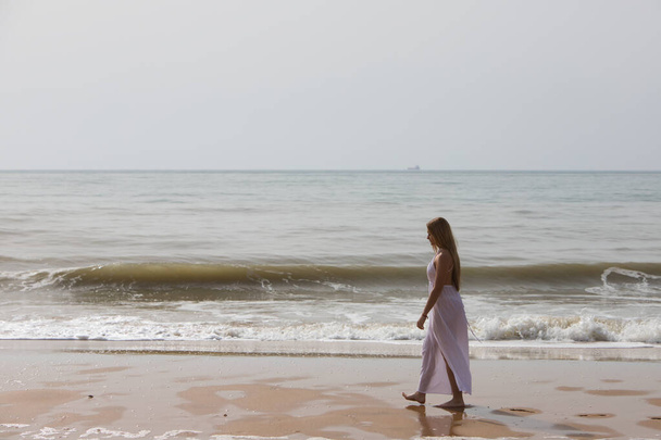 Jonge mooie blonde vrouw in witte jurk loopt op een zonnige dag over het zand aan de kust van het strand. De vrouw maakt verschillende lichaamsuitdrukkingen. Op de achtergrond de blauwe zee. - Foto, afbeelding