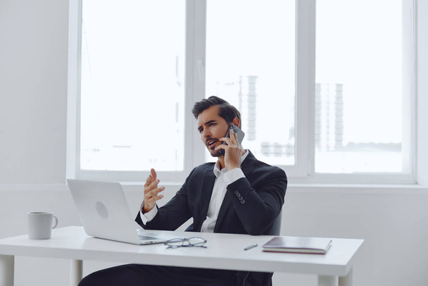 Человек в офисе бизнесмен сидит за столом и работает в ноутбуке и разговаривает по телефону, современный костюм, финансы и стартап концепция и анализ данных, рабочий образ жизни в ярком офисе. Высокое качество фото - Фото, изображение