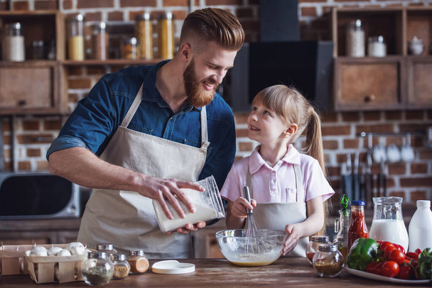 Милая маленькая девочка и ее красивый бородатый папа в фартуках взбивают яйца, добавляя муку и улыбаясь во время приготовления пищи на кухне
 - Фото, изображение