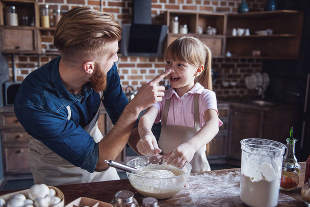 Cute little girl and her handsome bearded dad in aprons are having fun while baking in kitchen - Photo, Image
