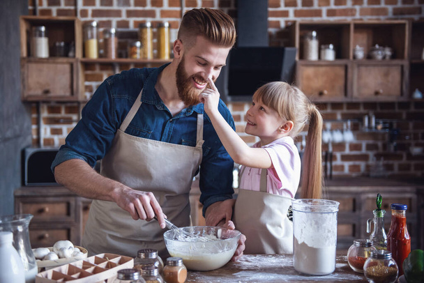 Милая маленькая девочка и ее красивый бородатый папа в фартуках готовят тесто и улыбаются во время приготовления пищи на кухне
 - Фото, изображение