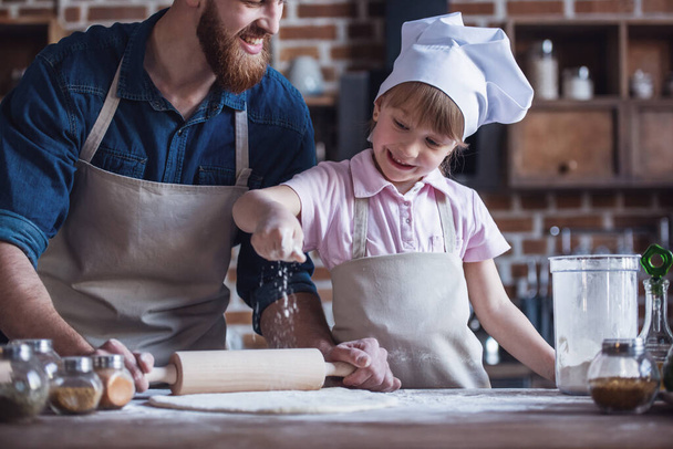 Schattig klein meisje en haar knappe bebaarde vader in schorten zijn afvlakken deeg met een deegroller en glimlachen tijdens het koken in de keuken - Foto, afbeelding