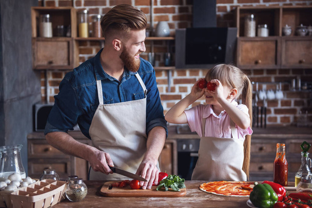 süßes kleines Mädchen und ihr hübscher bärtiger Vater in Schürzen lächeln beim Pizzakochen in der Küche - Foto, Bild
