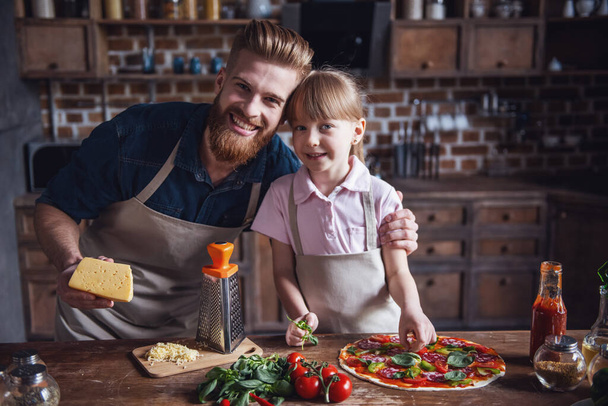 Aranyos kislány, kötények, szakállas, jóvágású apja kamera nézett és mosolyogva, miközben főzés pizza konyha - Fotó, kép