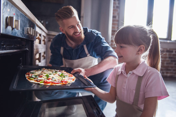 Carino bambina e il suo bel papà barbuto in grembiuli stanno parlando e sorridendo durante la cottura della pizza in cucina
 - Foto, immagini