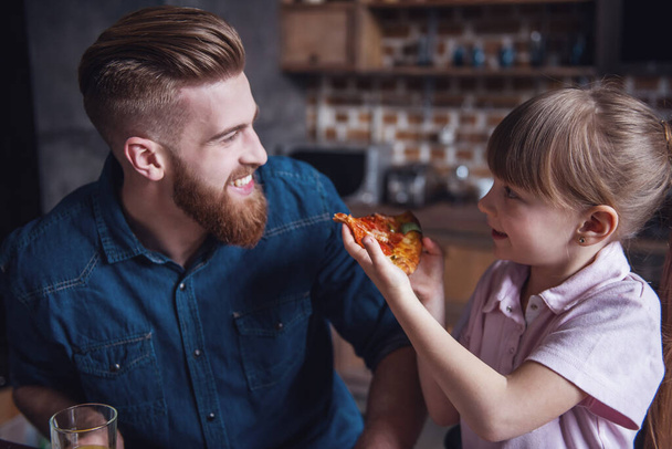 Aranyos kis lány és jóképű szakállas apja mosolyogva, miközben pizzát eszik a konyhában - Fotó, kép