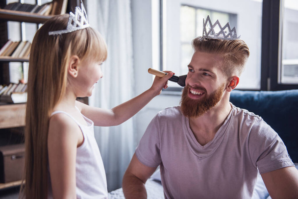 Мила маленька дівчинка і її красивий бородатий тато в коронках посміхаються, граючи в її кімнаті. Дівчина робить макіяж свого тата
 - Фото, зображення