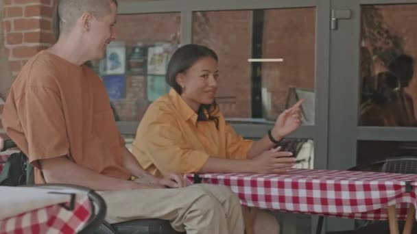 Mittlere Aufnahme von zwei kaukasischen Männern mit Behinderung und ihren fröhlichen Freundinnen, die sich bei einem Doppeldate im Sommercafé im Freien treffen - Filmmaterial, Video