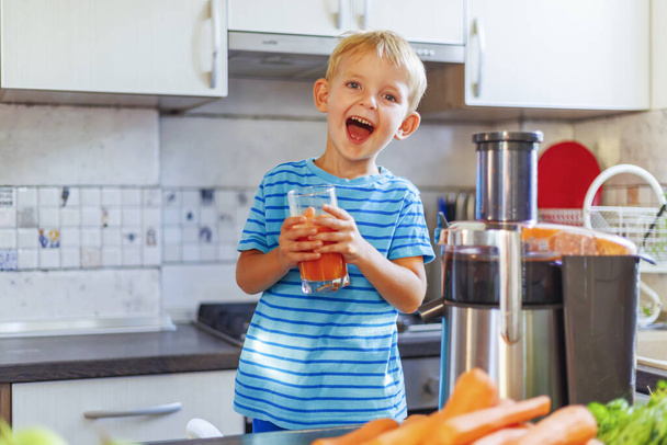 Szczęśliwe uśmiechnięte dziecko cieszące się świeżo wyciskanym sokiem marchewkowym w przytulnej kuchni w domu. Zdrowie i dobry nastrój przy prawidłowym odżywianiu - Zdjęcie, obraz