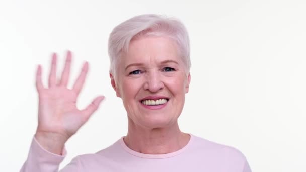 Vidám idős nő sugárzik meleg mosollyal, miközben integet kezét egy barátságos üdvözlés. A szeme szikrázik a kedvességtől és a gesztustól, valódi melegséget sugároz.. - Felvétel, videó