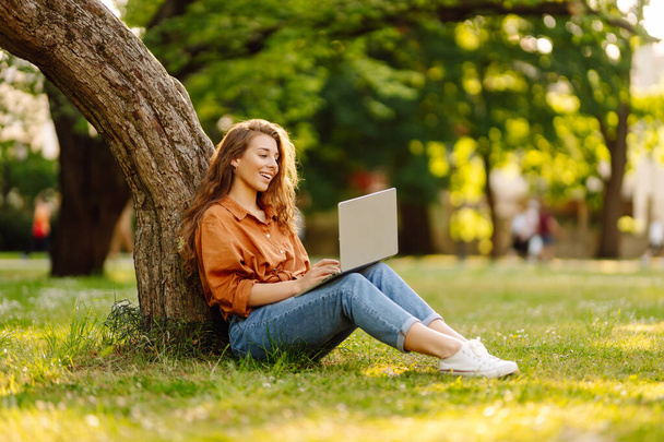 Happy woman sitting on a green clearing with a laptop. Portrait of a young freelance woman working outdoors. Online, technology concept. - Foto, immagini