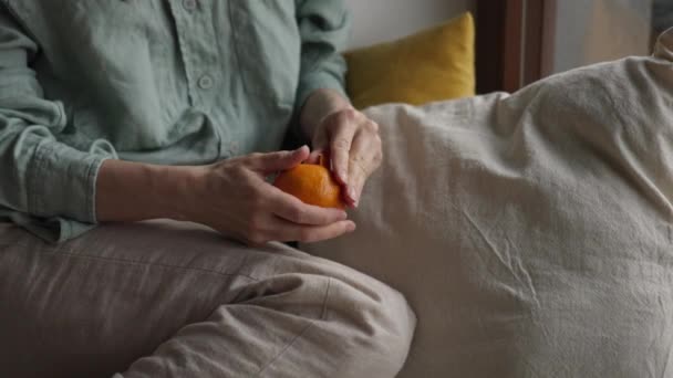 Close-up medium shot van meisje handen peeling mandarijn oranje fruit zitten op de vloer in de buurt van het raam in gezellige slaapkamer tijdens regenachtige trieste dag. Hoge kwaliteit 4k beeldmateriaal - Video