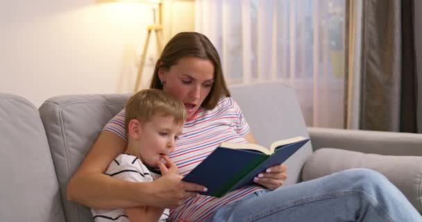 Μητέρα και γιος διαβάζουν ένα βιβλίο που κάθεται σε ένα άνετο καναπέ στο σαλόνι, ώρα για οικογένεια. Υψηλής ποιότητας 4k πλάνα - Πλάνα, βίντεο