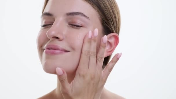 Woman gently applies face lotion, fingers gliding smoothly over skin with care and tenderness against a clean white backdrop. Her relaxed expression reflects the tranquility of the skincare ritual. - Footage, Video