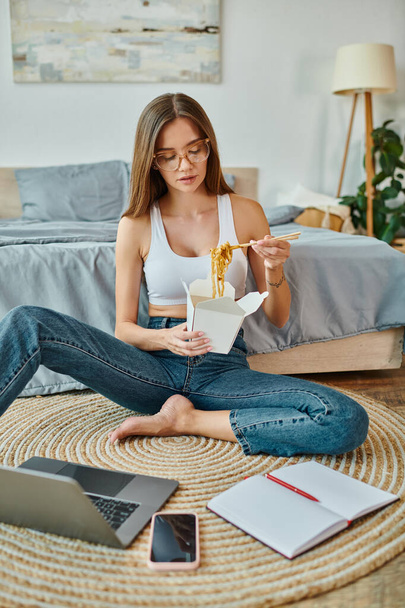 στοχαστική όμορφη γυναίκα σε ζεστό homewear τρώει noodles, ενώ εργάζονται εξ αποστάσεως σε φορητό υπολογιστή - Φωτογραφία, εικόνα