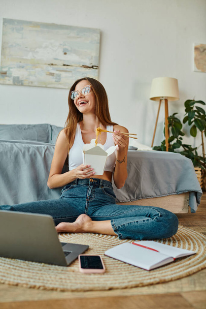 mujer atractiva alegre con gafas en ropa de casa disfrutando de fideos mientras trabaja desde casa de forma remota - Foto, imagen