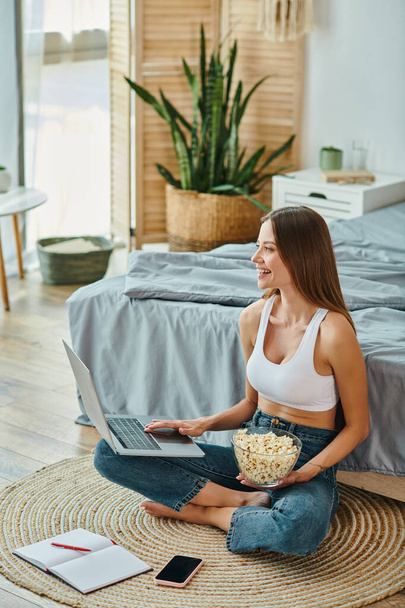 joyeuse femme attrayante en vêtements de maison assis sur le sol avec du pop-corn tout en travaillant à distance sur un ordinateur portable - Photo, image