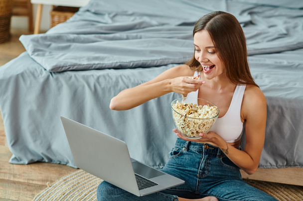 atractiva mujer sentada en el suelo con las piernas cruzadas comiendo palomitas de maíz y viendo películas en el portátil - Foto, imagen