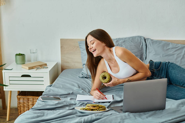 vrolijk aantrekkelijke vrouw in homewear met lang haar eten heerlijke donut tijdens het werken van thuis - Foto, afbeelding