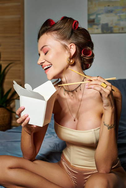 χαρούμενη ελκυστική γυναίκα με μπούκλες μαλλιών απολαμβάνοντας νόστιμα noodles από το κουτί, ενώ κάθεται στο κρεβάτι - Φωτογραφία, εικόνα