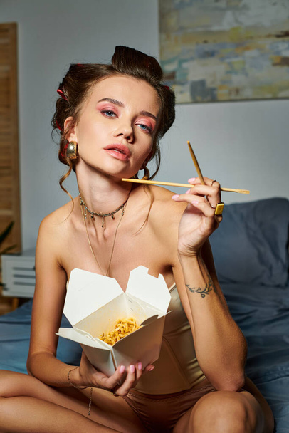 attraktive junge Frau mit beigem Korsett auf dem Bett sitzend mit einer Schachtel Nudeln und Essstäbchen in der Hand - Foto, Bild