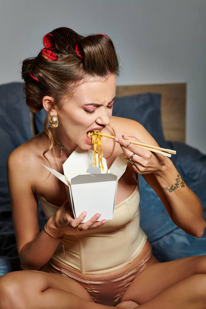 aantrekkelijke jonge vrouw in sexy beige korset eten noedels met eetstokjes terwijl zitten op bed - Foto, afbeelding