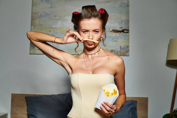 csinos fiatal nő szexi bézs corsete a haj curlers pózol sült krumplival nézi kamera - Fotó, kép