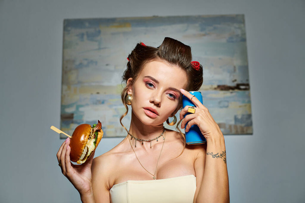 atraente mulher sexy no espartilho bege com encrespadores de cabelo posando com hambúrguer e refrigerante olhando para a câmera - Foto, Imagem