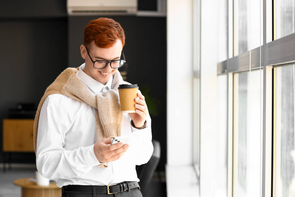 Молодой владелец бизнеса с чашкой кофе с помощью мобильного телефона в офисе - Фото, изображение