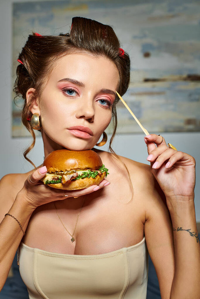 красивая привлекательная женщина в бежевом сексуальном корсете держит в руках огромный вкусный бургер и смотрит в камеру - Фото, изображение