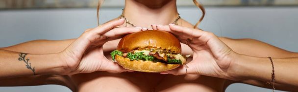 abgeschnittene Ansicht einer jungen Frau mit Accessoires und Tätowierung auf der Hand, die riesige Burger zeigt, Banner - Foto, Bild