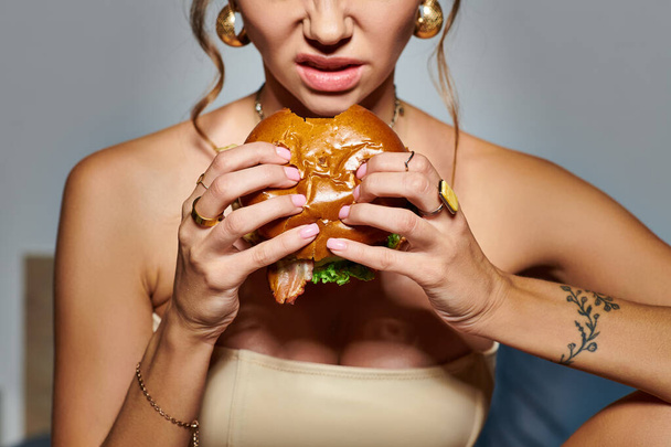 巨大なハンバーガーを食べる手のアクセサリーとタトゥーを持つ感情的な若い女性のクロップビュー - 写真・画像