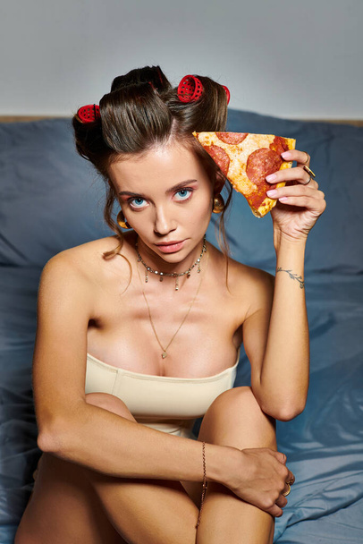 atrakcyjna kobieta z lokówkami i akcesoriami pozującymi z kawałkiem pizzy i patrzących w kamerę - Zdjęcie, obraz