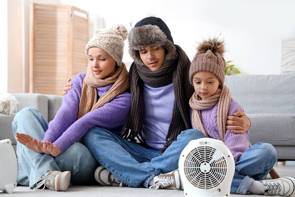 Κατεψυγμένα οικογένεια σε χειμερινά ρούχα θέρμανση κοντά σε ηλεκτρικούς θερμαντήρες ανεμιστήρα στο σπίτι - Φωτογραφία, εικόνα
