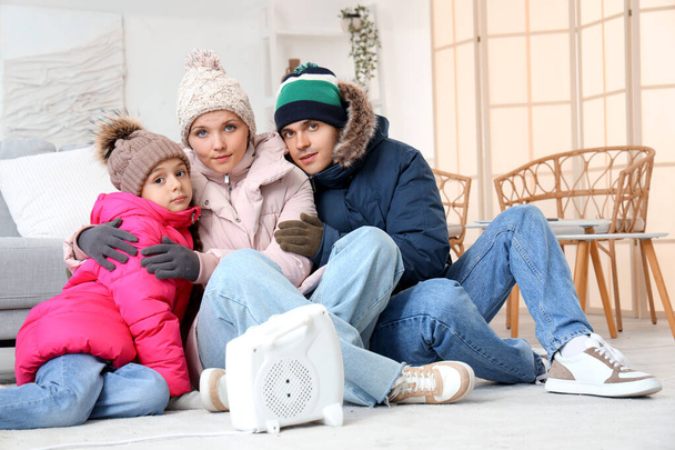 Evde elektrikli ısıtıcının yanında kışlık giysiler içinde donmuş bir aile ısınıyor. - Fotoğraf, Görsel