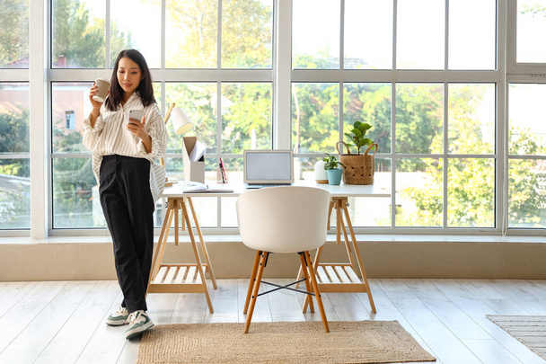 Mujer asiática joven con taza de café y teléfono móvil en la oficina en casa - Foto, imagen