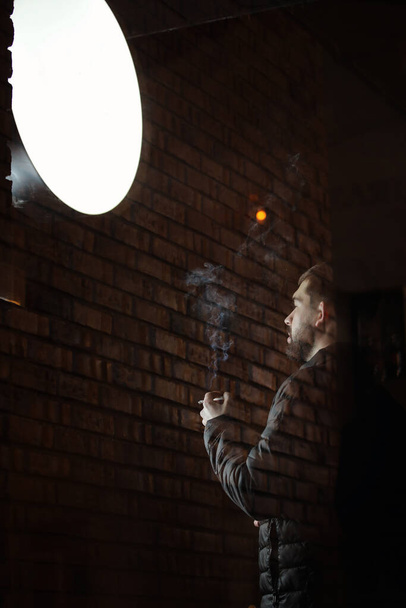 Muž s plnovousem si užívá kouření venku po náročném dnu za soumraku u cihlové zdi. za skleněnými dveřmi. Cigarety pomáhají se vším od nudy po zvládání hněvu - Fotografie, Obrázek