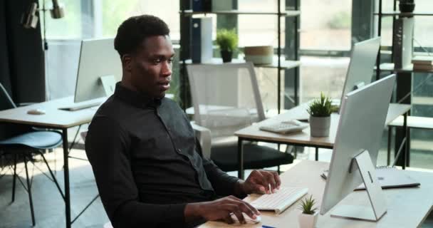 Afro-Amerikaanse man zit aan een bureau in het kantoor, typend op een toetsenbord met een warme glimlach. Zijn positieve en productieve houding straalt door de werkplek en creëert een aangename sfeer.. - Video