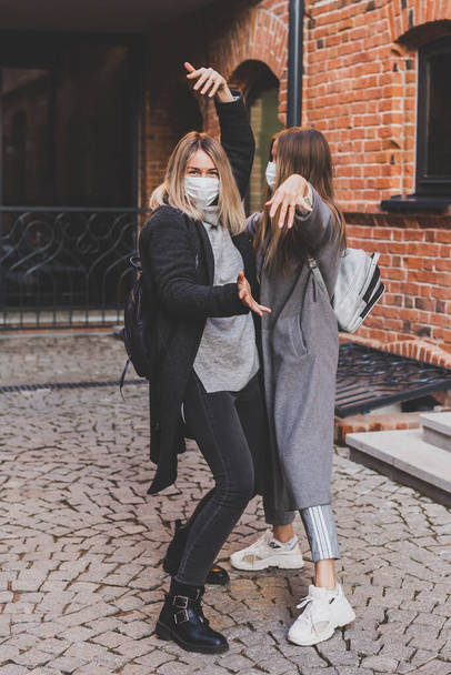 Dwie młode stylowe kobiety w maskach medycznych rozmawiające i śmiejące się podczas spaceru ulicą po mieście. Przyjaciele spacery na świeżym powietrzu w mieście - Zdjęcie, obraz