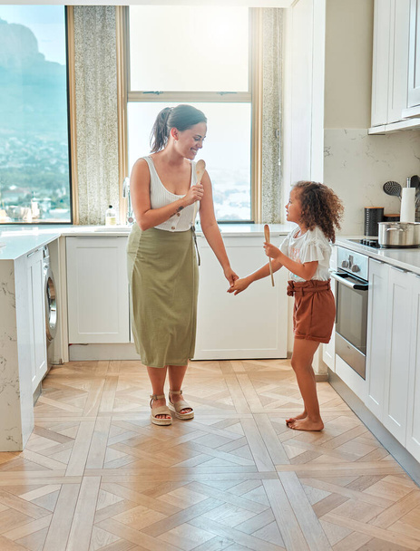 Mother and little daughter singing and dancing in the kitchen. Mixed race mom and child holding wooden spoons while having fun at home. - Фото, изображение
