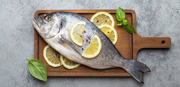 Nyers főzés nélküli hal dorádó citrommal és friss bazsalikom fa vágódeszkán késsel rusztikus kő háttér felülnézet, főzés egészséges hal dorádó koncepció - Fotó, kép