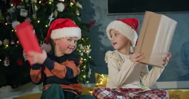 Τα παιδιά του νηπιαγωγείου κουνάνε τα χριστουγεννιάτικα δώρα ενάντια στο διακοσμημένο δέντρο με γιρλάντα. Τα αδέρφια απολαμβάνουν να γιορτάζουν μαζί την Πρωτοχρονιά στο σπίτι. - Πλάνα, βίντεο