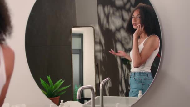 Afro-Amerikalı bir kadın banyoya girer ve arkadaşlarıyla mutlu bir şekilde konuşur. Etnik kız ayna yansımasına bakar saç stili kıvırcık saçlı, akıllı telefondan konuşur. - Video, Çekim